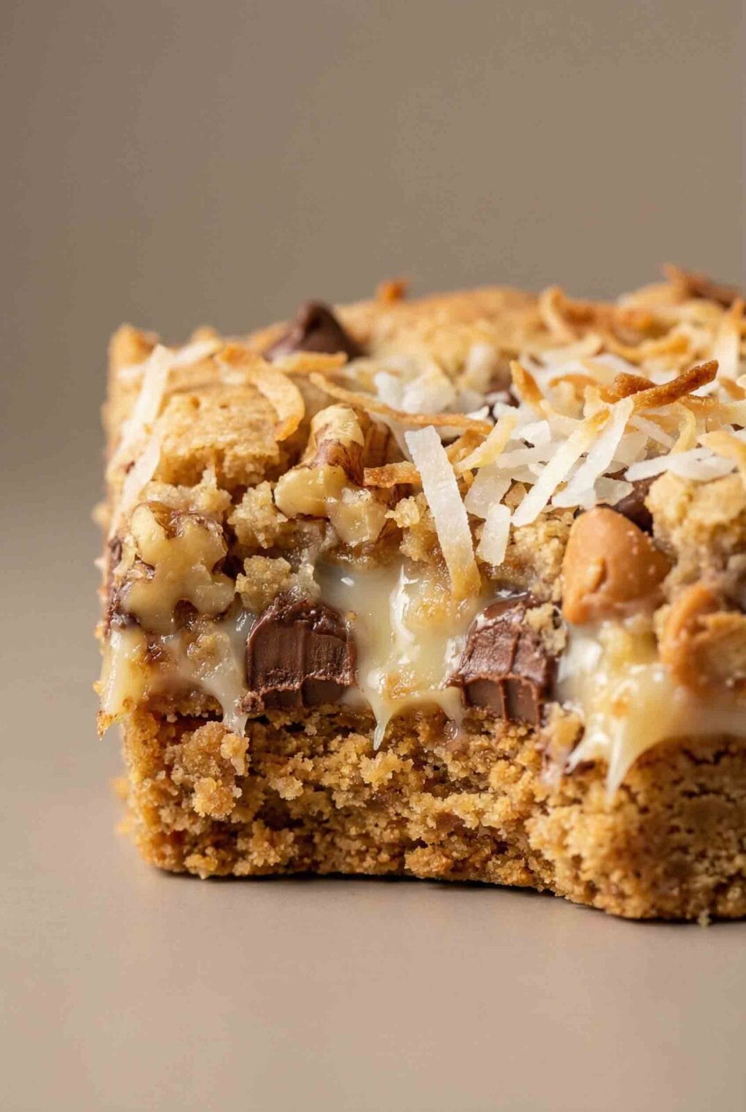 Close-up vertical macro photo of a 7 layer magic cookie bar showing detailed graham crust, intact chocolate and butterscotch chips, nuts, coconut shreds, and condensed milk texture on a smooth warm gray background.