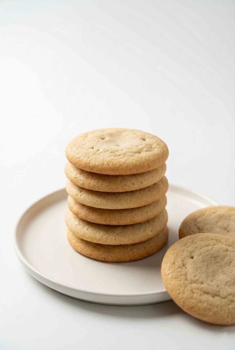 Stack of classic sugar cookies on a matte white ceramic plate in a clean white studio setting with soft cinematic lighting.