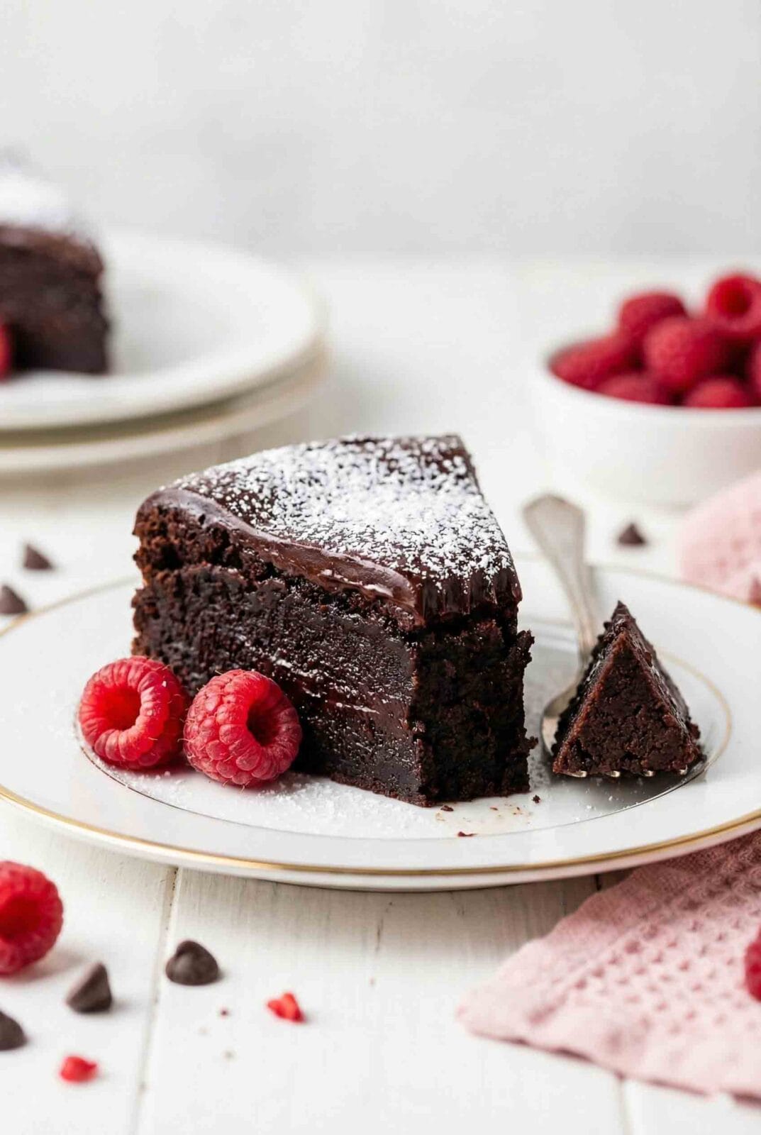 Slice of flourless chocolate cake gluten free with dense fudgy texture, chocolate ganache topping, powdered sugar, and fresh raspberries on white plate with gold rim