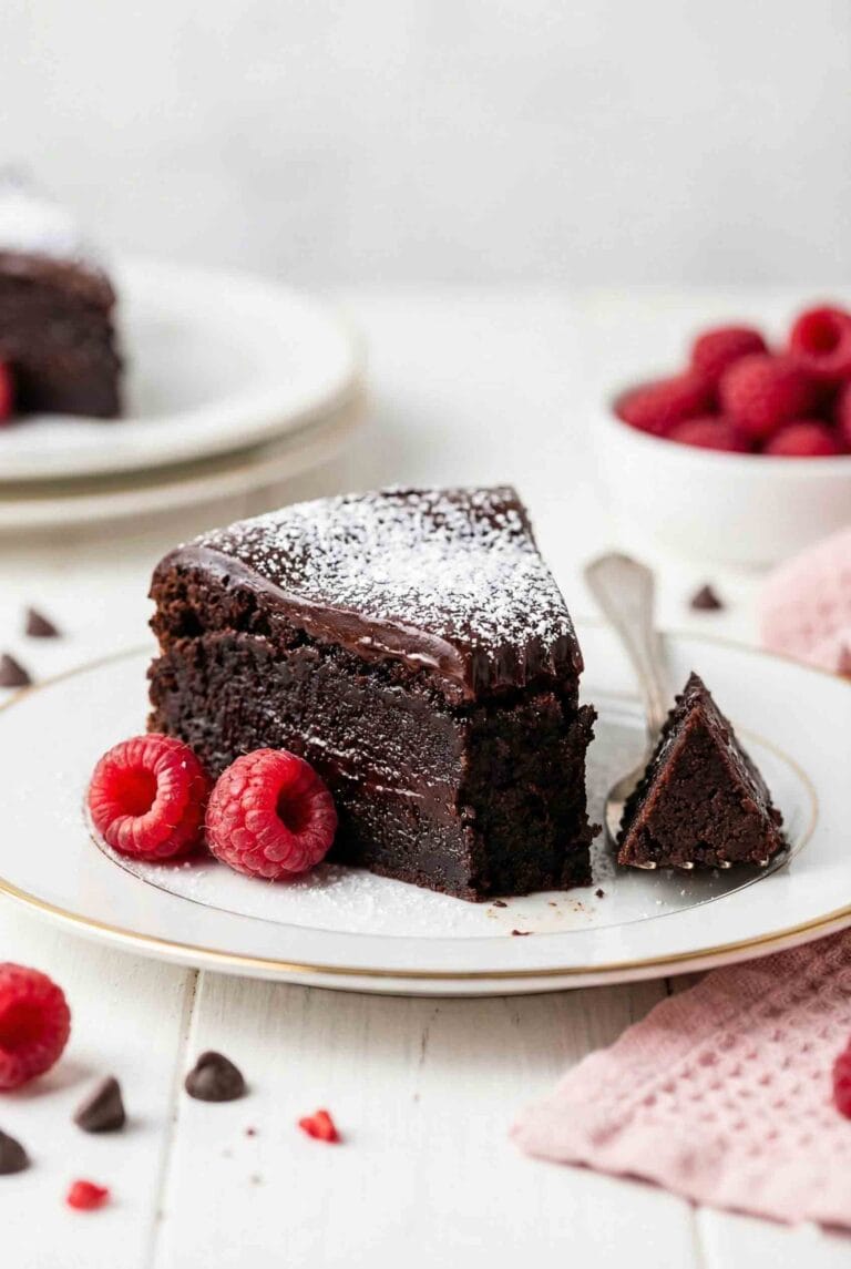 Slice of flourless chocolate cake gluten free with dense fudgy texture, chocolate ganache topping, powdered sugar, and fresh raspberries on white plate with gold rim