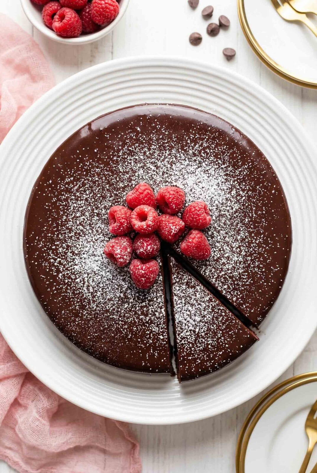 Overhead view of whole flourless chocolate cake gluten free in white dish with glossy chocolate ganache, powdered sugar dusting, and fresh raspberries on top with slice cut out