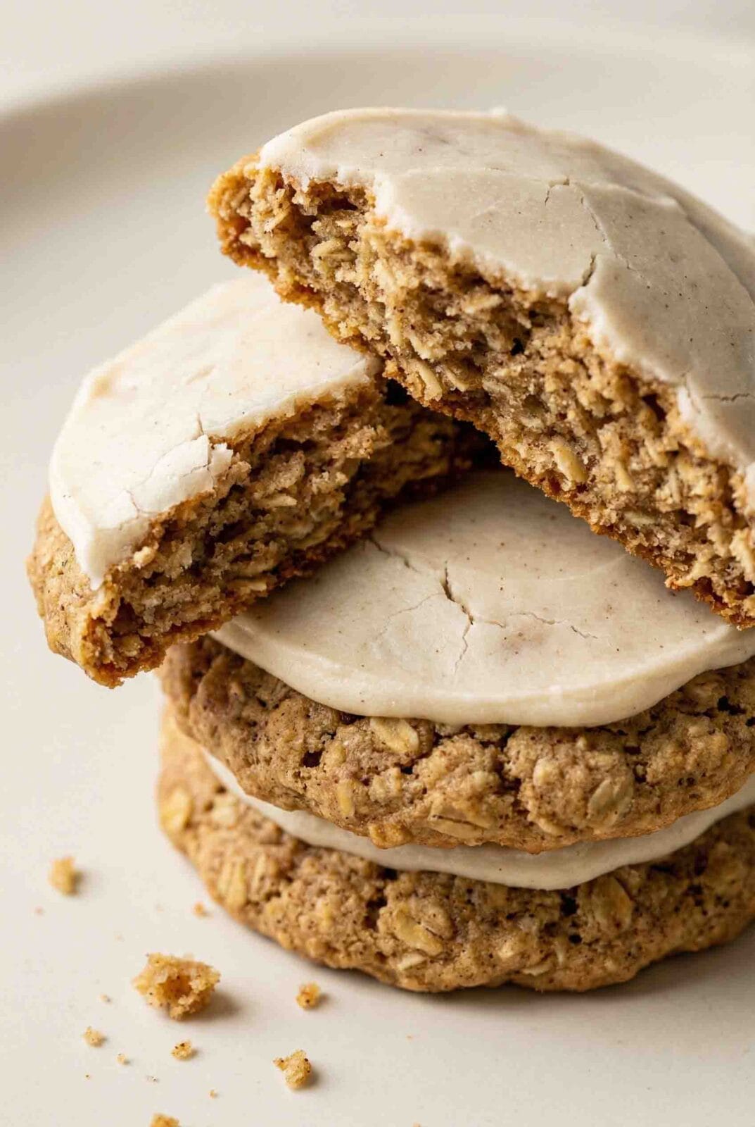 Extreme macro photo of stacked iced oatmeal cookies with the top cookie broken to reveal the detailed oat crumb interior and hardened vanilla icing, captured on a warm soft-white background with diffused natural light.