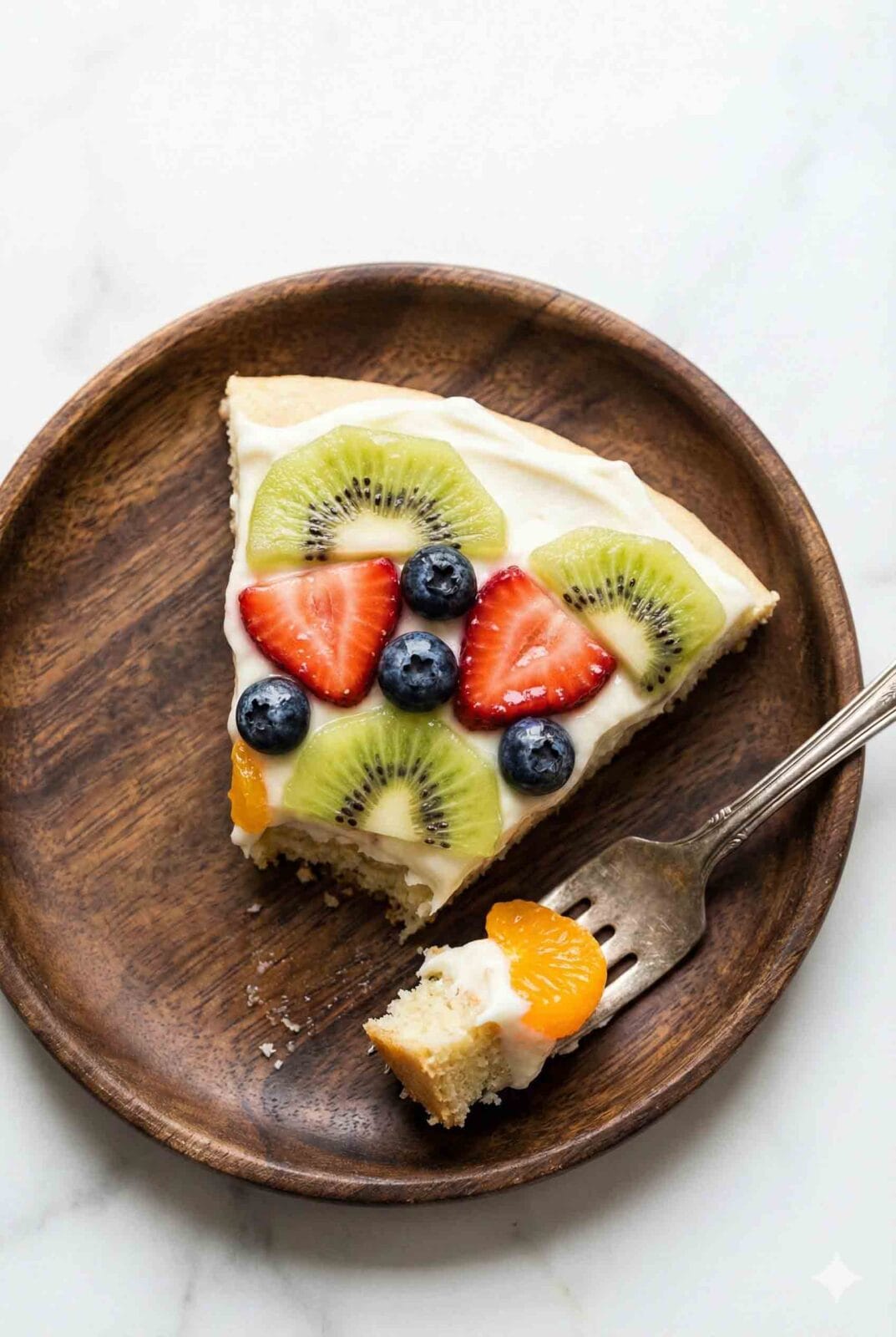 A 45-degree macro close-up of a fruit pizza slice on a white ceramic plate in a minimalist studio setting, topped with kiwi, strawberries, and blueberries.
