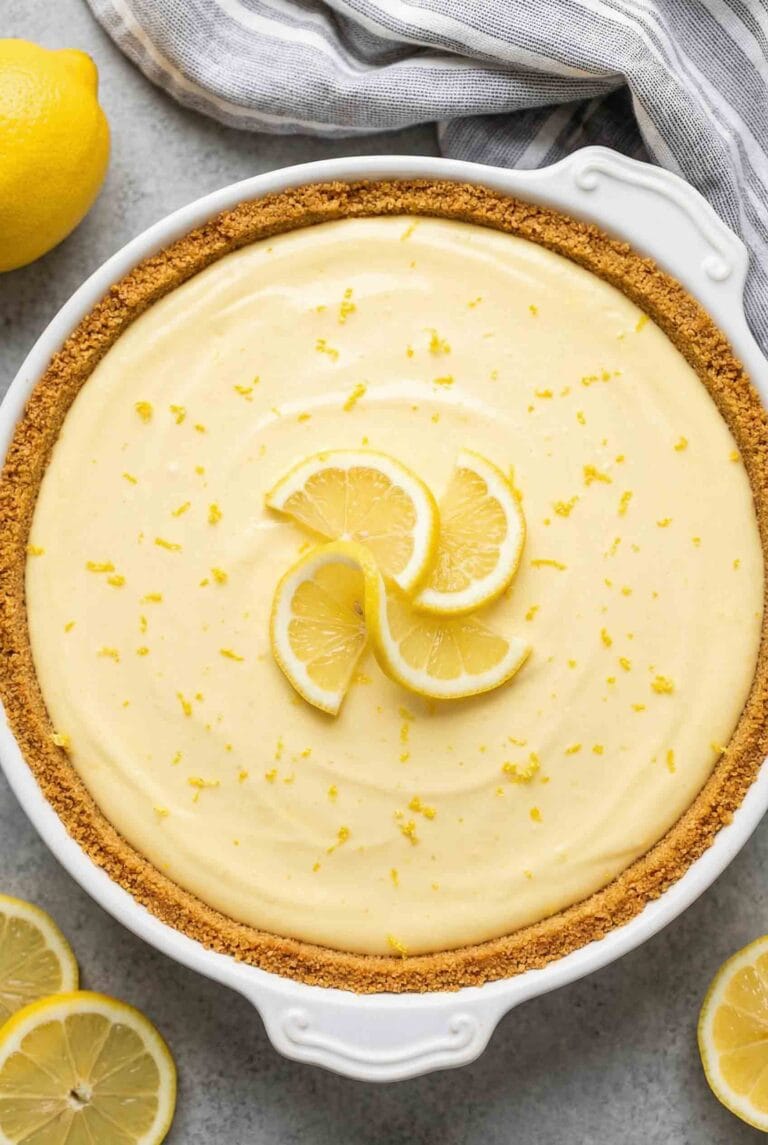 Overhead view of whole no-bake lemon pie in white ceramic dish with smooth pale yellow filling, graham cracker crust, topped with lemon slices and zest, surrounded by fresh lemons