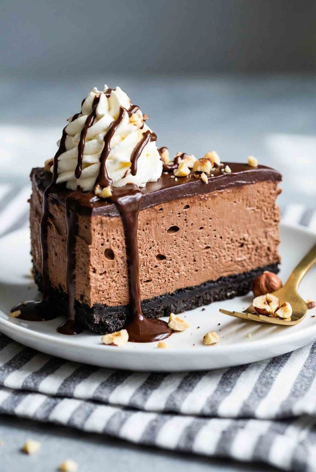 Tall slice of no-bake Nutella cheesecake with airy chocolate filling, dark cookie crust, whipped cream swirl, Nutella drizzle, and hazelnuts
