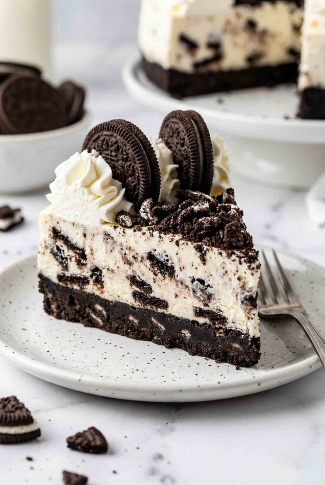Close-up slice of no-bake Oreo cheesecake on white plate showing layers of Oreo crust, creamy cheesecake filling with cookie chunks, and whipped cream topping with whole Oreos and fork