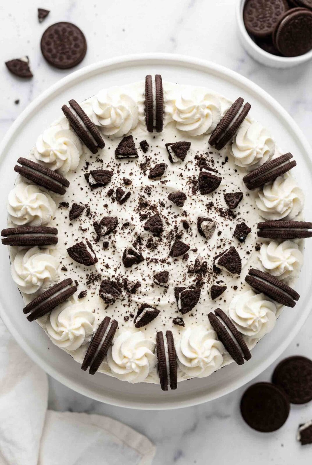 Overhead view of whole no-bake Oreo cheesecake decorated with whipped cream rosettes, whole Oreo cookies around border, and crushed cookie pieces on white plate with scattered Oreos