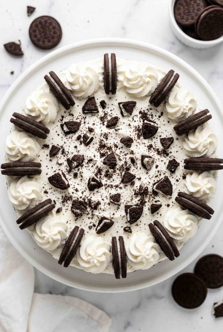 Overhead view of whole no-bake Oreo cheesecake decorated with whipped cream rosettes, whole Oreo cookies around border, and crushed cookie pieces on white plate with scattered Oreos