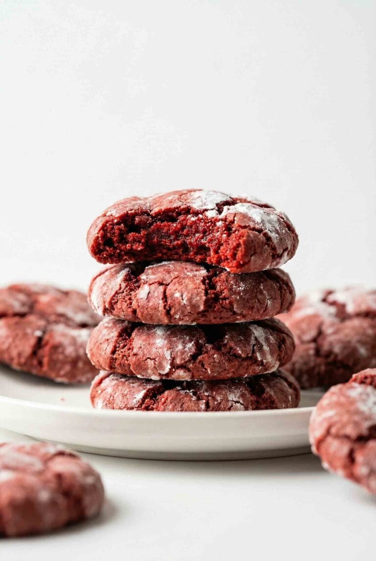 Close-up 45° studio image of four stacked red velvet crinkle cookies on a white plate, top cookie bitten, with detailed powdered-sugar texture and clean white backdrop.