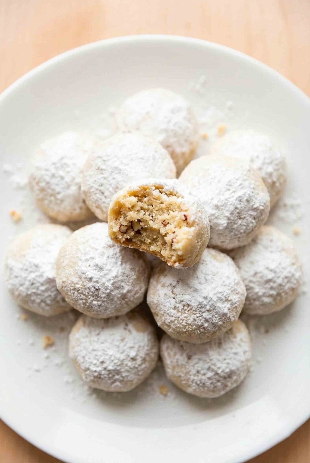 Eight snowball cookies dusted with powdered sugar on a matte white plate with almonds in a clean white studio setting