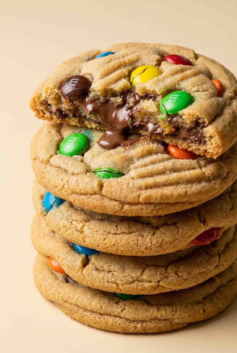 Thick and soft M&M cookies stacked high with colorful candy pieces and gooey chocolate centers, photographed in a clean, modern food photography style